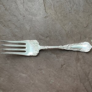 May include: A silver fork with an ornate handle. The fork has four tines and is on a gray background.