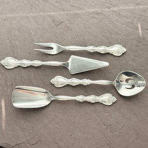 Può includere: Set di posate argentate tra cui una forchetta, una paletta per torte, un cucchiaio da portata e un cucchiaio. Gli utensili hanno manici decorati e sono disposti su una superficie grigio scuro. Il set è per servire il cibo.