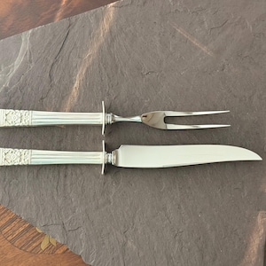 Könnte beinhalten: Ein silbernes Tranchierbesteck, bestehend aus einem Messer und einer zweizinkigen Gabel, liegt auf einer dunkelgrauen Schieferoberfläche. Die Griffe weisen ein dekoratives Blumendesign auf. Das Set ist zum Tranchieren und Servieren von Fleisch bestimmt.