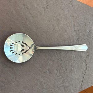 May include: Silver slotted spoon with a decorative design on the bowl. The spoon has a long handle with a floral pattern near the end. The bowl features a stylized design with cut-out shapes.