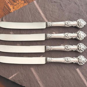 May include: Four silver butter knives with decorative handles and rounded blades are displayed on a dark grey surface. The handles feature a floral design, adding elegance to the cutlery set. The knives are suitable for serving butter or spreads.