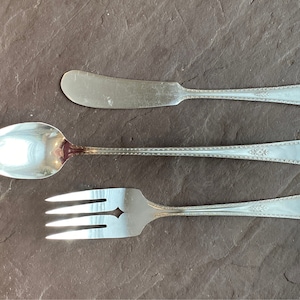 Puede incluir: Un juego de tres cubiertos plateados: un cuchillo para mantequilla, una cuchara y un tenedor. Los cubiertos tienen un patrón decorativo en los mangos.