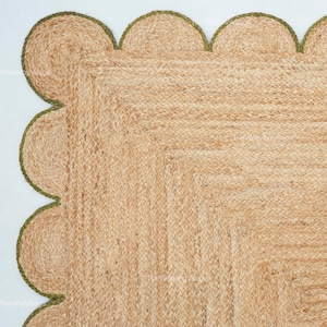 May include: A rectangular jute rug with a scalloped edge. The rug is a natural tan color with a green border. The weave is visible, and the rug appears to be handmade. The rug is on a white background.