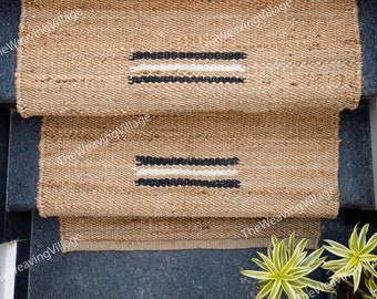 May include: Natural jute stair treads with a rectangular design. Each tread features a horizontal stripe pattern in black and white. The treads are placed on dark gray steps, with a glimpse of a white wall and greenery in the background.