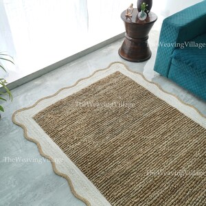 May include: A rectangular jute rug with a scalloped edge. The rug is a natural brown colour with a cream-coloured border. The rug is placed on a grey concrete floor. The Weaving Village is printed on the rug.