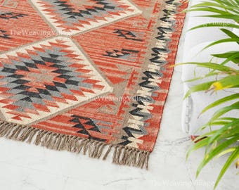 May include: A rectangular, hand-woven rug with a geometric diamond pattern in shades of red, grey, black, and beige. The rug has a fringed edge and is placed on a white marble floor. The rug's design is a mix of tribal and modern styles.