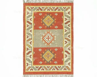 May include: Rectangular rug with a geometric design in shades of red, grey, yellow, and cream. The rug features a central diamond pattern and a fringed edge. The Weaving Village is printed on the rug.