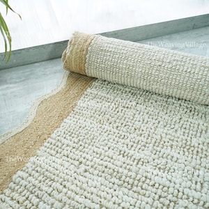 May include: A rolled-up, cream-coloured rug with a textured, looped pile. The rug has a beige border along one edge and is partially unrolled, revealing the textured surface. The text "TheWeavingVillage" is visible in the image.