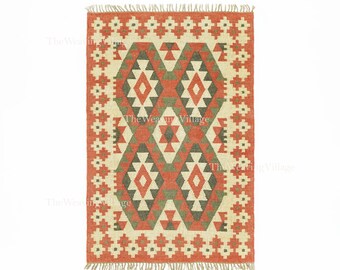 May include: Rectangular rug with a geometric pattern in red, cream, and green. The rug has a central diamond design and a border with repeating floral motifs. It has a fringed edge and is suitable for a living room or bedroom. The rug's dimensions are approximately 120 cm x 180 cm.