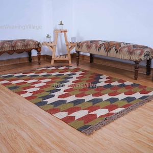 Op de afbeelding: Een rechthoekig vloerkleed met een geometrisch patroon in rood, groen, wit en grijs. Het vloerkleed heeft een gefranjerde rand en ligt op een houten vloer. Twee banken en een kleine houten plantenstandaard staan op de achtergrond.