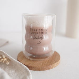 Könnte beinhalten: Eine doppelwandige Glas-Tasse mit einem braunen Getränk. Auf der Tasse steht "Einatmen Ausatmen Lächeln" und ein Smiley.