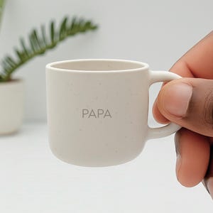 Könnte beinhalten: Eine kleine, cremefarbene Keramiktasse mit dem Wort "PAPA" auf der Vorderseite. Die Tasse hat eine abgerundete Form und einen einzelnen Henkel. Die Tasse wird von einer Person gehalten.