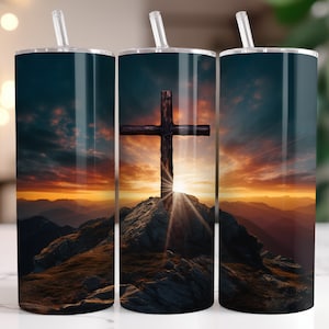 Könnte beinhalten: Ein Edelstahlbecher mit einem Sonnenuntergangs- und einem Holzkreuz-Design auf einem Berggipfel. Der Becher wird mit einem Strohhalm und einem Deckel geliefert.