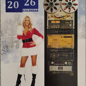 Op de afbeelding: Een 2026 wandkalender met een vrouw in een kerstman outfit en een stapel bandrecorders. De kalender heeft het nummer 12 en de woorden "REEL TO REEL RECORDERS" in het rood.