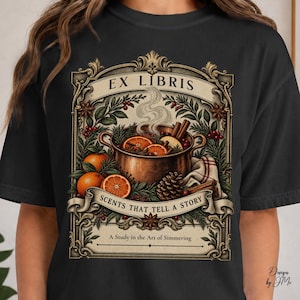 May include: A black t-shirt featuring a graphic of a copper pot filled with simmering oranges, cinnamon sticks, and pinecones. The design includes the text "EX LIBRIS," "SCENTS THAT TELL A STORY," and "A Study in the Art of Simmering" in a frame.