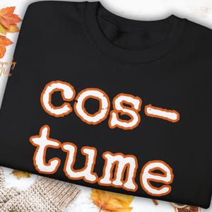May include: Black sweatshirt with the word "COSTUME" in white letters with orange trim. The sweatshirt is surrounded by fall leaves and a knitted sweater.