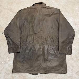 Op de afbeelding: Bruine jas met lange mouwen, een knoopkraag en een trekkoord in de taille. De jas heeft een versleten uiterlijk en een klassiek ontwerp, geschikt voor buitenactiviteiten. De jas is gemaakt van duurzaam materiaal.