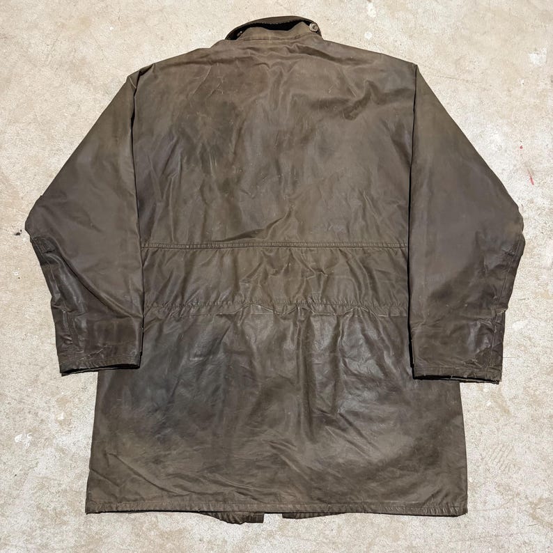 Op de afbeelding: Bruine jas met lange mouwen, een knoopkraag en een trekkoord in de taille. De jas heeft een versleten uiterlijk en een klassiek ontwerp, geschikt voor buitenactiviteiten. De jas is gemaakt van duurzaam materiaal.