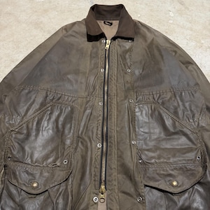 Op de afbeelding: Een bruine gewaxte katoenen jas met een donkerbruine corduroy kraag. De jas heeft een lange ritssluiting, drukknoopsluitingen en twee grote voorzakken met kleppen. De jas is een vintage stijl.