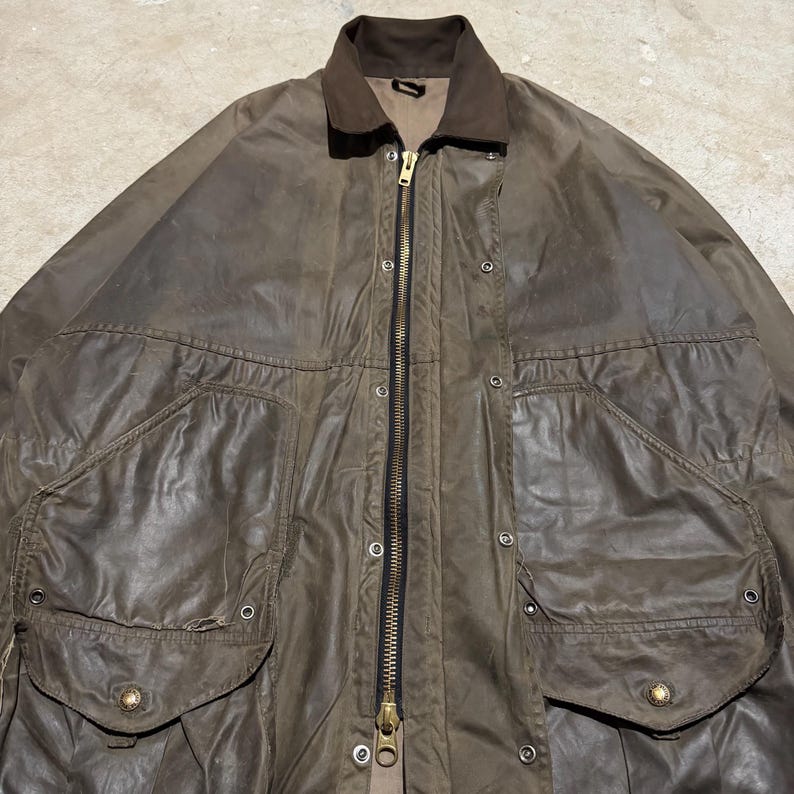 Op de afbeelding: Een bruine gewaxte katoenen jas met een donkerbruine corduroy kraag. De jas heeft een lange ritssluiting, drukknoopsluitingen en twee grote voorzakken met kleppen. De jas is een vintage stijl.