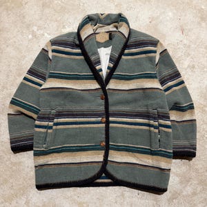 Puede incluir: Una chaqueta de manga larga con cuello tipo chal y un patrón de rayas horizontales en tonos verde salvia, beige, azul oscuro y negro. La chaqueta incluye tres botones marrones en la parte delantera, dos bolsillos laterales y un forro interior blanco.