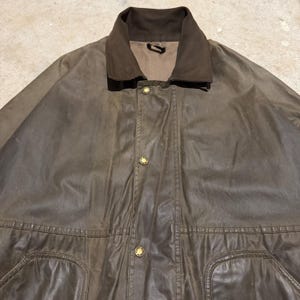 Op de afbeelding: Bruine gewaxte katoenen jas met een corduroy kraag en goudkleurige knopen. De jas heeft twee voorzakken en een klassiek ontwerp, geschikt voor buitenactiviteiten.