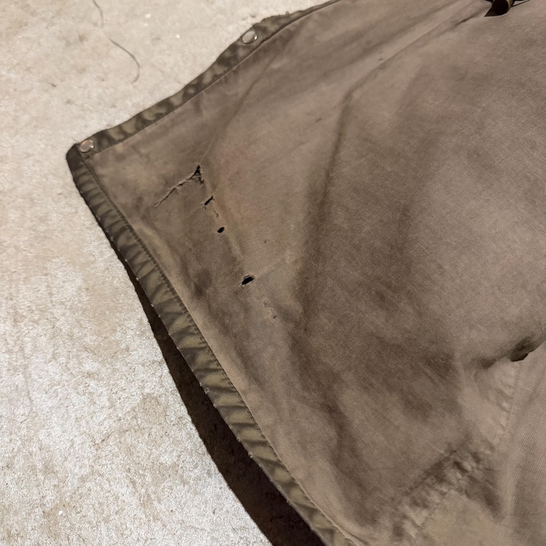 Op de afbeelding: Close-up van een versleten, olijfgroen canvas materiaal met zichtbare slijtage. De stof heeft verschillende kleine gaatjes en een gestikte rand. De randen zijn versterkt met een donkergroene bies en metalen drukknopen zijn zichtbaar.
