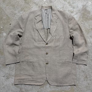 May include: A beige blazer featuring a three-button front closure, notched lapels, and two lower welt pockets. The interior lining visible at the collar displays a vertical white and grey pinstripe pattern. The garment has long sleeves and a structured fit.