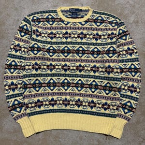 May include: A long-sleeve crewneck sweater by Polo by Ralph Lauren featuring a yellow base with a repeating geometric fair isle pattern in shades of blue, dark red, and white. The garment has a ribbed collar, cuffs, and hem in solid yellow.