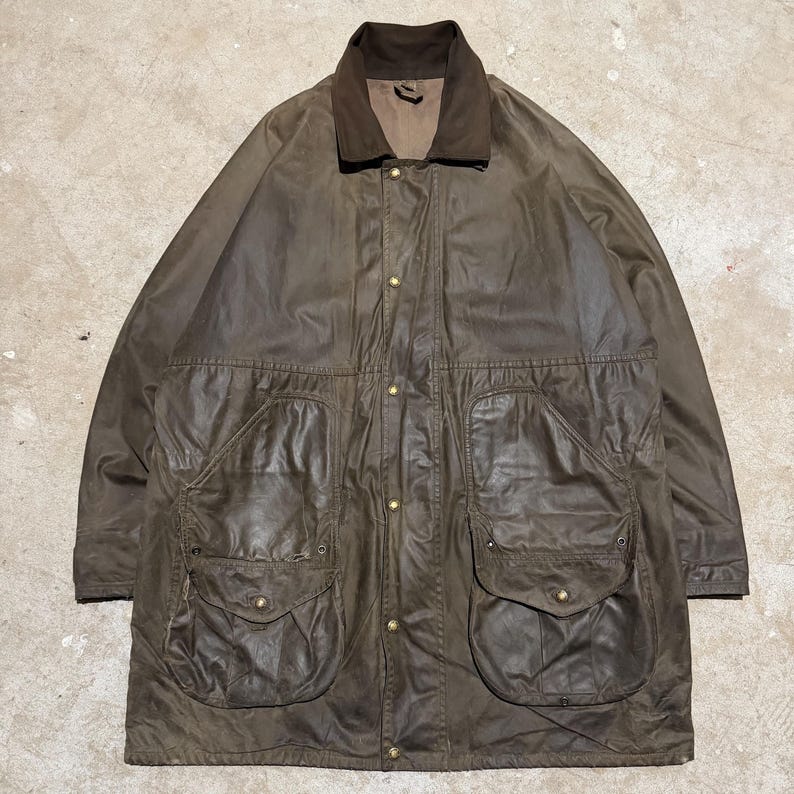 Op de afbeelding: Bruine gewaxte katoenen jas met een corduroy kraag en vier grote voorzakken. De jas heeft een knoopsluiting en een relaxte pasvorm, geschikt voor buitenactiviteiten. De jas heeft een gedempte bruine kleur.