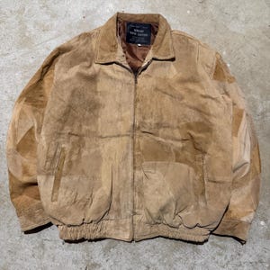 Può includere: Giacca in pelle scamosciata marrone con un design patchwork. La giacca ha un collo, una chiusura con cerniera e due tasche anteriori. L'orlo inferiore e i polsini sono elasticizzati. La giacca è etichettata "GENUINE SUEDE LEATHER".