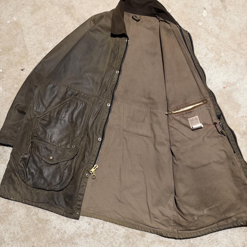 Op de afbeelding: Een bruine gewaxte katoenen jas met een corduroy kraag, open om een beige voering en binnenzak te laten zien. De jas heeft twee grote voorzakken met drukknoopsluitingen en een messing ritssluiting.