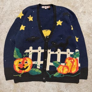 Puede incluir: Cárdigan azul marino con diseño de Halloween. Presenta una valla blanca, calabazas, estrellas, murciélagos y un gato negro. El cárdigan tiene cierre de botones y ribete negro.