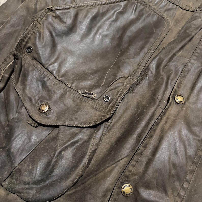 Op de afbeelding: Close-up van een versleten, bruine gewaxte katoenen jas. Kenmerken zijn een grote zak met klep, metalen drukkers en een verweerde uitstraling. De jas heeft een vintage esthetiek.