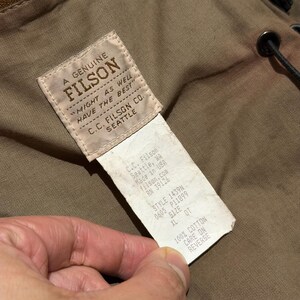 Op de afbeelding: Close-up van een Filson-label, met de merknaam en de tekst "MIGHT AS WELL HAVE THE BEST." Het label bevat ook het adres van C.C. Filson Co. in Seattle, WA, en productdetails zoals stijl en maat XL.