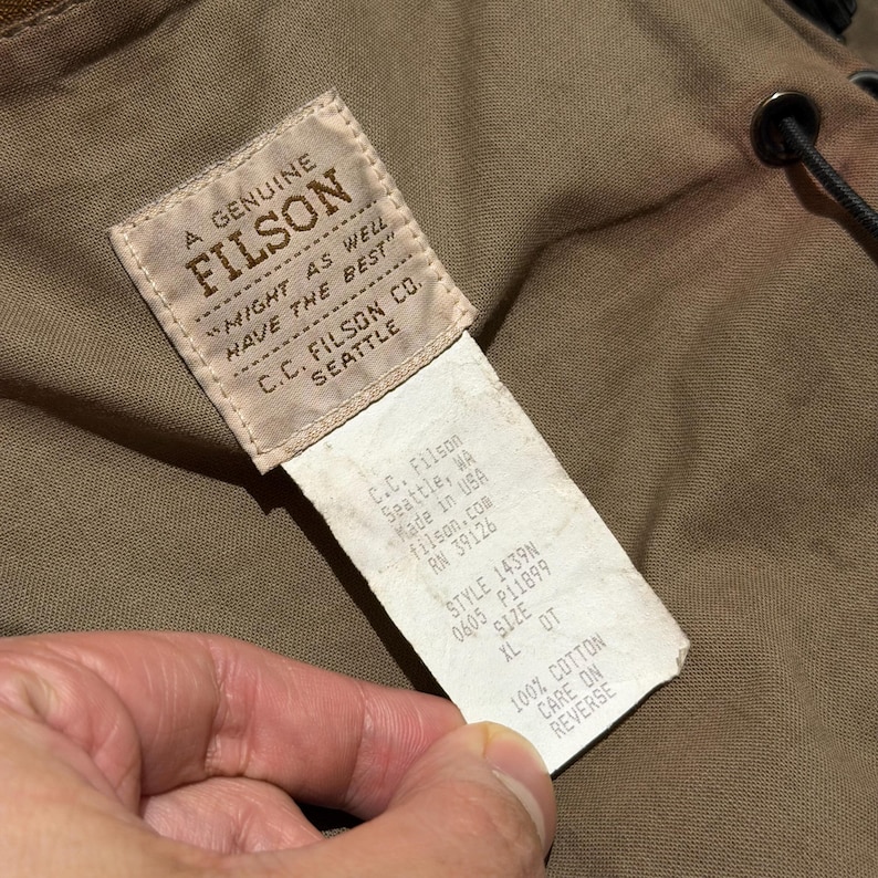Op de afbeelding: Close-up van een Filson-label, met de merknaam en de tekst "MIGHT AS WELL HAVE THE BEST." Het label bevat ook het adres van C.C. Filson Co. in Seattle, WA, en productdetails zoals stijl en maat XL.