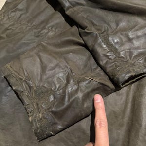 Op de afbeelding: Close-up van een bruine jasmouw met zichtbare slijtage. De stof lijkt gewaxt of behandeld, met een licht glanzende afwerking. Stiksels en naden zijn zichtbaar, wat duidt op leeftijd en gebruik. De mouw is gevouwen.