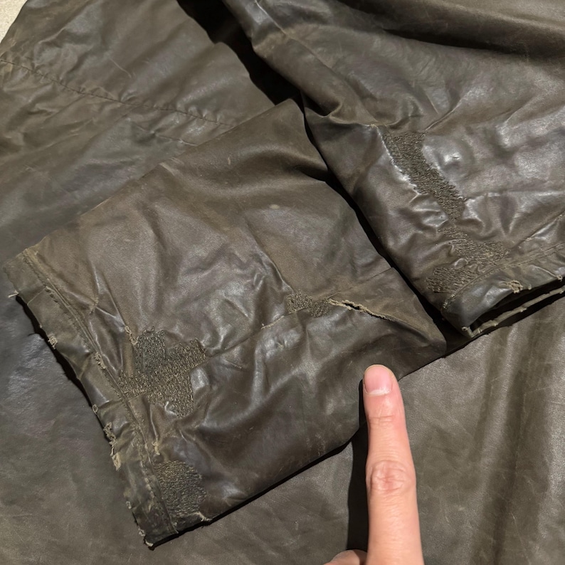 Op de afbeelding: Close-up van een bruine jasmouw met zichtbare slijtage. De stof lijkt gewaxt of behandeld, met een licht glanzende afwerking. Stiksels en naden zijn zichtbaar, wat duidt op leeftijd en gebruik. De mouw is gevouwen.