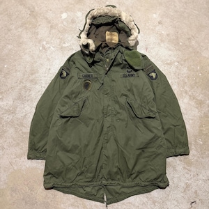 May include: An olive green military-style parka with a fur-lined hood. The jacket features multiple pockets, patches, and the text "U.S. Army" and "Gardner". The parka is designed for cold weather and has a button and zipper closure.