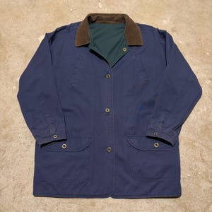 May include: A navy blue jacket featuring a brown corduroy collar and a dark green inner lining. The garment includes a front button closure with metallic buttons, two large lower pockets with buttoned flaps, and long sleeves with buttoned cuffs.