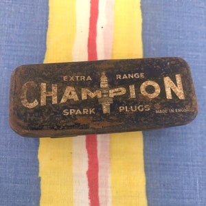 Può includere: Una scatola di metallo arrugginita con le parole "EXTRA RANGE CHAMPION SPARK PLUGS MADE IN ENGLAND" stampate su di essa. La scatola è di colore marrone scuro con un'etichetta gialla e bianca sbiadita.