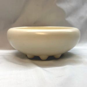 May include: A cream-colored ceramic bowl with a smooth, rounded rim and a wide, shallow basin. The bowl is supported by three small, rounded feet evenly spaced around the base, providing a stable foundation for the vessel.