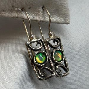 May include: A pair of silver dangle earrings featuring a rectangular frame with intricate scrollwork and a central round cabochon stone displaying iridescent green and orange flashes of color, suspended from curved silver ear wires.