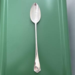 May include: A silver spoon with an oval bowl and a long, slender handle. The spoon is made of a shiny, reflective metal and is set against a green background. The spoon's handle has a decorative, flared end.