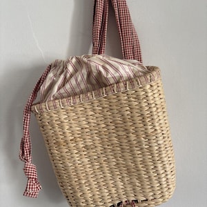 Puede incluir: Un bolso de mimbre con cierre de cordón. El bolso presenta un color beige natural con un forro interior a rayas rojas y blancas. El asa y el cordón están hechos de tela a cuadros rojos y blancos.
