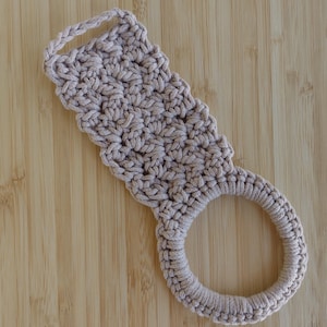 May include: A beige, handmade crochet kitchen towel holder with a loop for hanging. The holder features a textured, rectangular top section and a circular ring at the bottom. The item is displayed on a wooden surface.