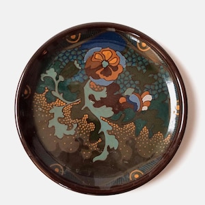 May include: A round, decorative plate with a dark brown rim. The plate features a floral design with orange, blue, and green hues. The center of the plate has a detailed flower with intricate leaf patterns and small orange dots.