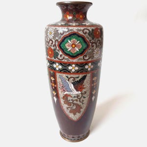May include: A tall, cylindrical cloisonné vase featuring a dark brown base color with intricate, multi-colored enamel patterns. The design includes a central flying bird motif, floral arrangements in red, white, and green, and decorative geometric borders.