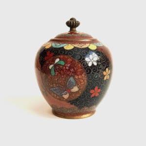 Pode incluir: Um pote redondo com tampa apresentando um trabalho intrincado de esmalte cloisonné. A superfície exibe um fundo preto com padrões de redemoinho, acentuado por motivos florais vermelhos, amarelos e azuis e um design de borboleta num círculo.