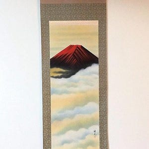 May include: A traditional Japanese scroll painting depicting a red mountain peak rising above a sea of white clouds. The mountain is painted in a stylised manner with dark lines and a red hue. The clouds are painted in a soft, ethereal style with shades of white, blue, and yellow. The scroll is mounted on a brown silk backing with a green border.